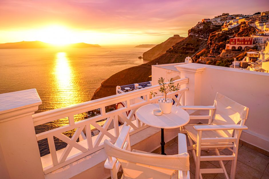 Idyllic Santorini sunset from a patio overlooking the caldera, with white chairs and table. – How do the two islands compare side by side?