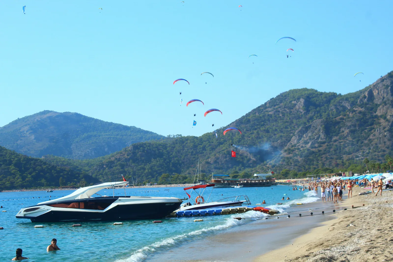 Multiple paragliders landing above the crowded beach at Oludeniz, Turkey, with a boat and Turkish flag smoke trail below