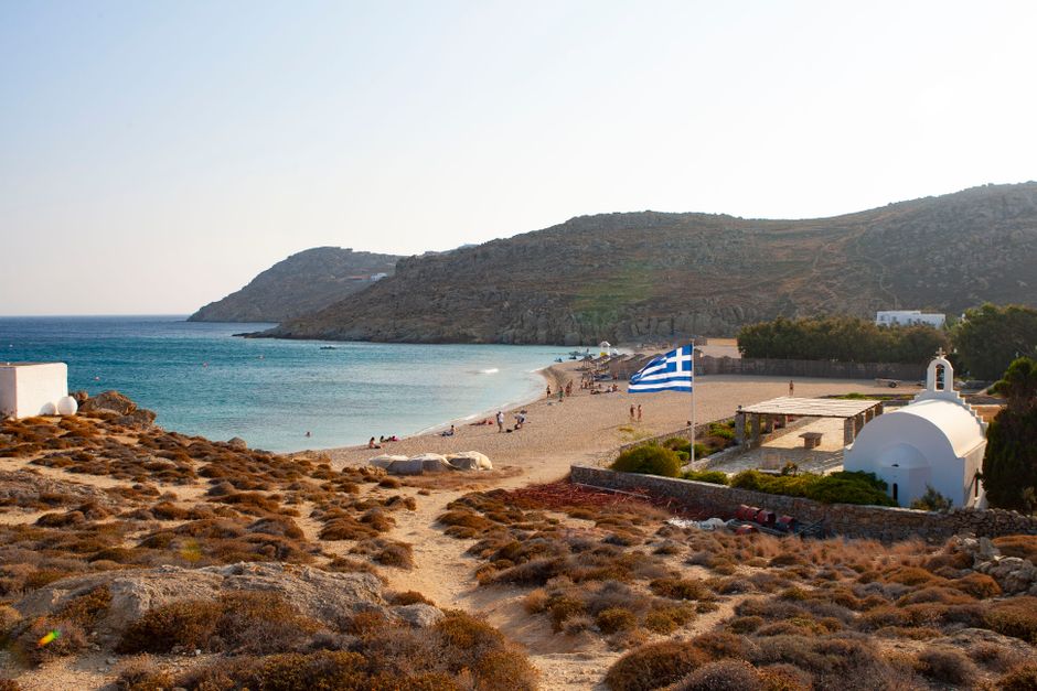 Scenic view of a beach in Mykonos with a Greek flag and traditional church. – Mykonos: Elia, Agrari & Fokos — Cyclades Nudism Without the Rulebook
