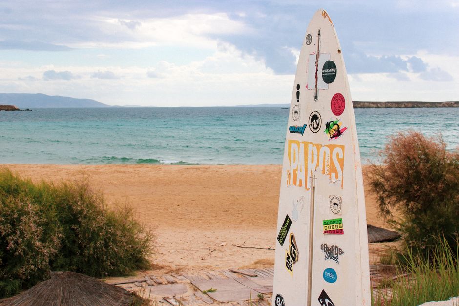A surfboard with stickers on Paros beach with cloudy skies and a tranquil sea. – Paros: Lageri Beach, the Cyclades' Quiet Naturist Retreat