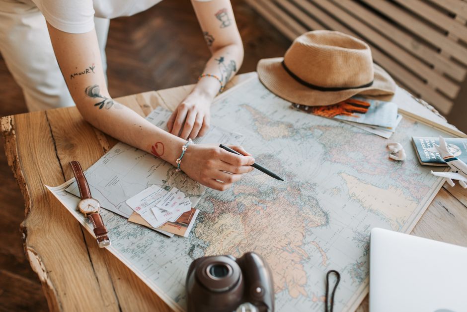 Tattooed woman planning a trip with a world map, passports, and travel essentials on a wooden table. β 2. Read the Price Graph Before You Lock In Dates