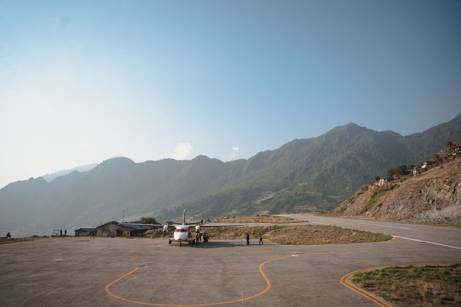 A scenic view of a small aircraft on a mountain airstrip surrounded by lush green hills under a vibrant blue sky. – The Classic Route: Lukla to Base Camp Day by