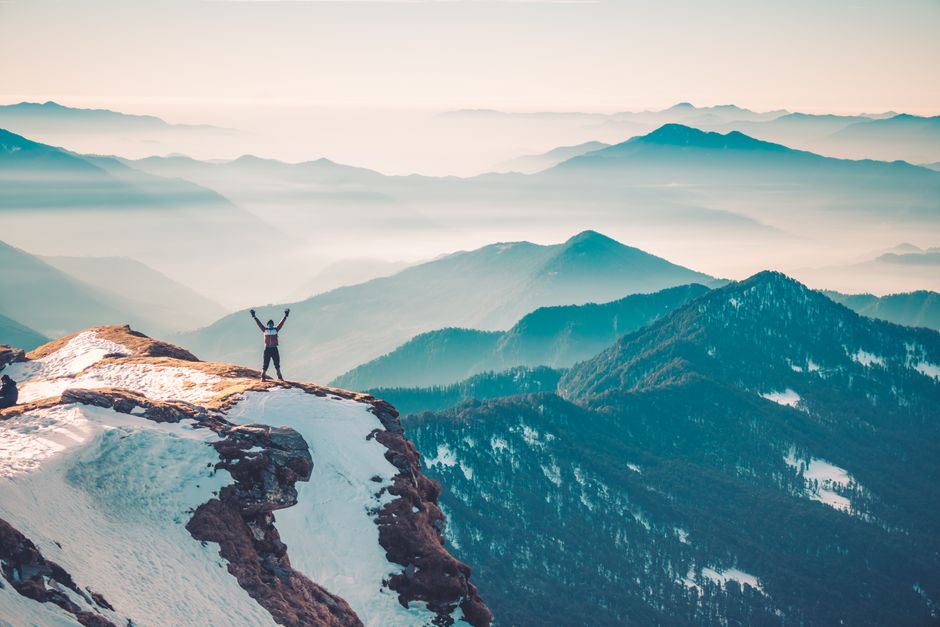 A hiker stands on a snowy mountain peak, arms raised in triumph, surrounded by a vast mountain range. – Summit Success Rates: Why More Days Beats a Cheaper Pack