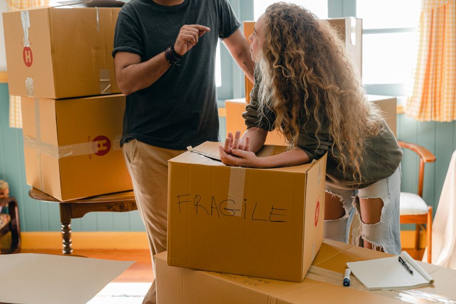 Crop faceless man in wristwatch talking to girlfriend in fashion jeans with wavy hair while standing near box with FRAGILE note after moving into new house – Re