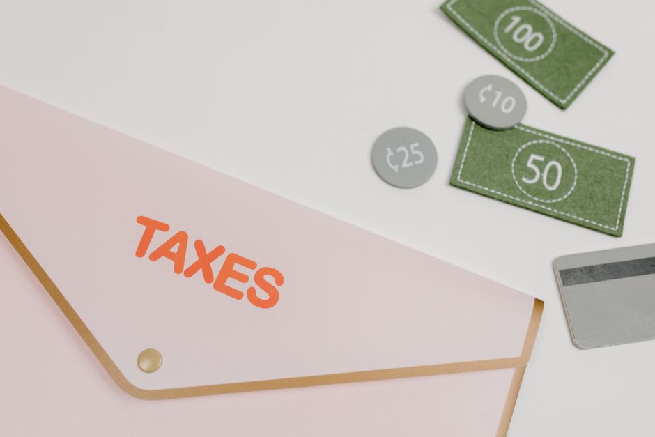 Pink envelope labeled 'TAXES' with play money and card on white background. – Do You Still Pay Tax Back Home?