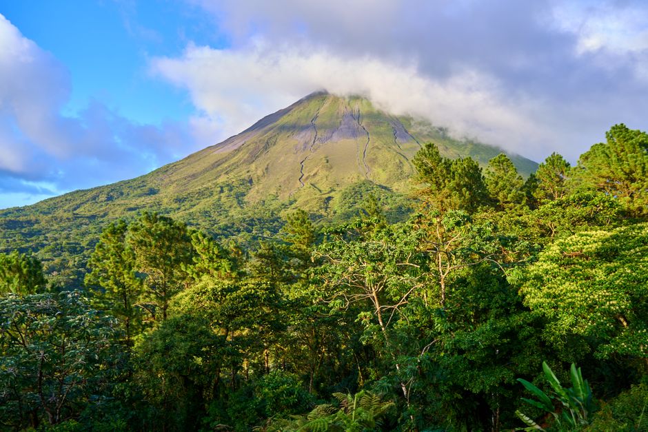 Lush green forest with a prominent volcanic mountain under a blue sky with clouds. – Arenal Volcano Zipline Tours: Views and Adrenaline
