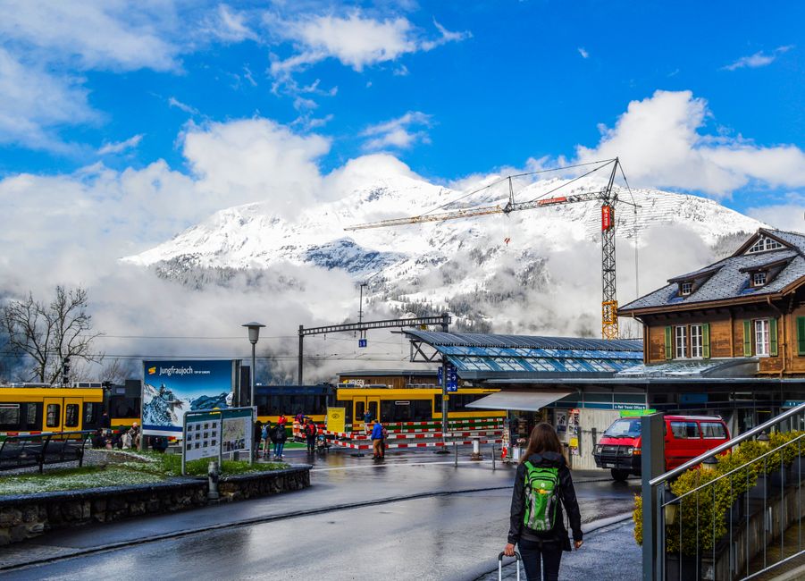 Back view of anonymous female with suitcase walking on street to modern train station in city near snowy mountains in sunny day – Getting to Interlaken: Flights