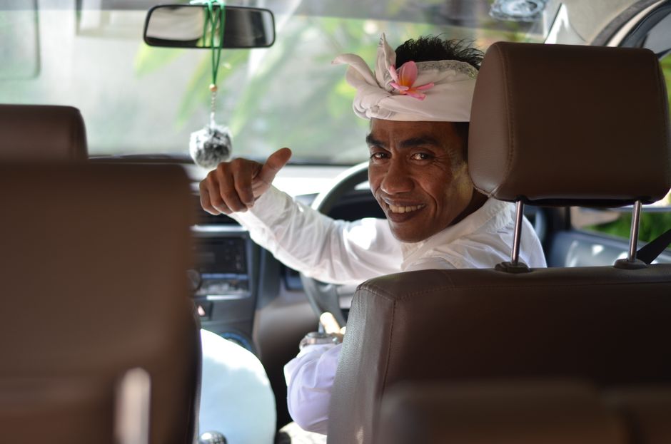 A Balinese man wearing traditional attire gives a thumbs up and smiles warmly from inside a car in Ubud, Bali. – Getting There Without the Stress