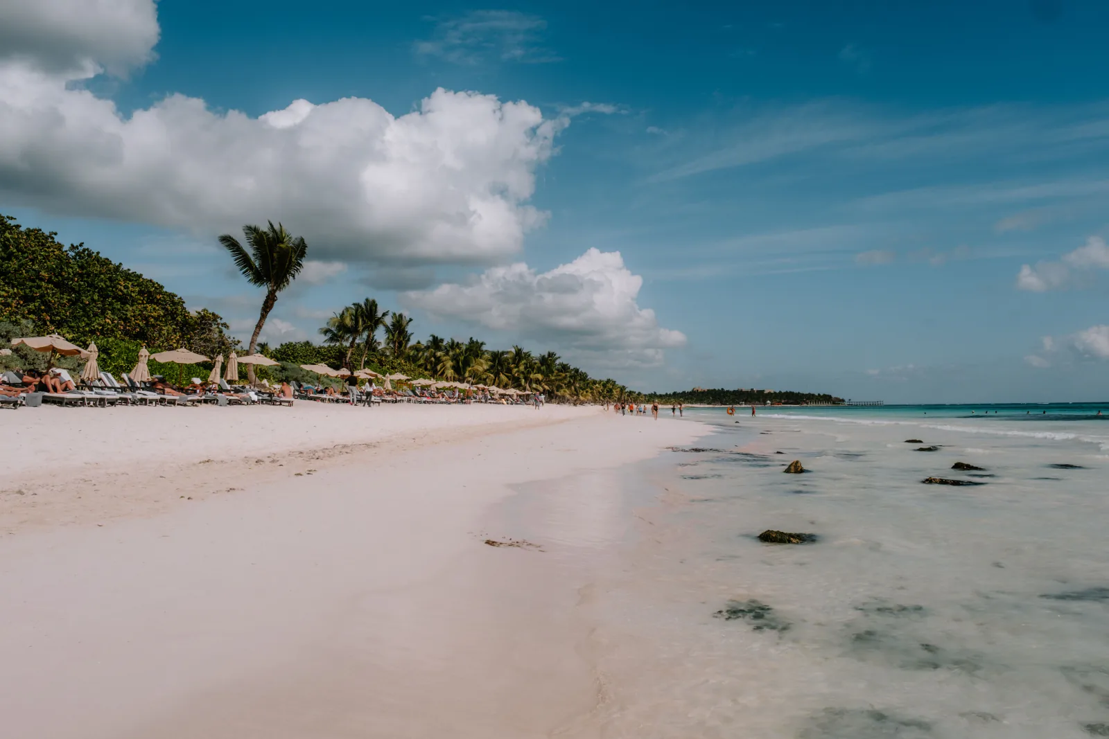 Serene beach in Tulum, Mexico with palm trees, clear water, and sunbathers under a blue sky. – Zamna Tulum vs Tulum's Other Beach Day Parties