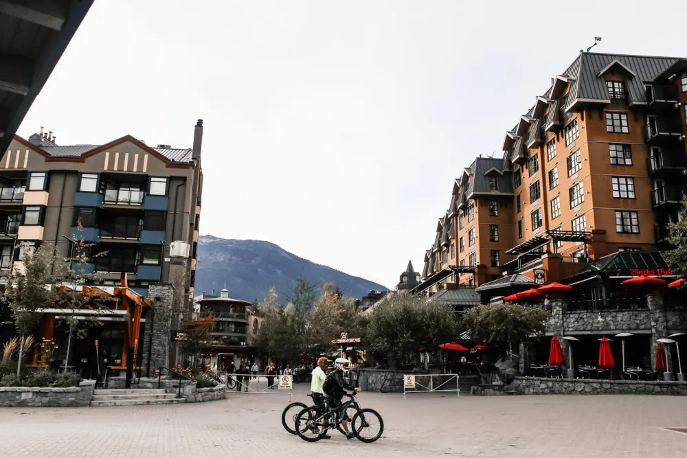 Cyclists walking their bikes across Whistler Village square – what a Whistler mountain biking trip costs, BC