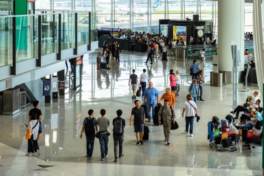Travellers walking through a glass-walled terminal concourse – weighing separate-ticket savings, Hong Kong International Airport