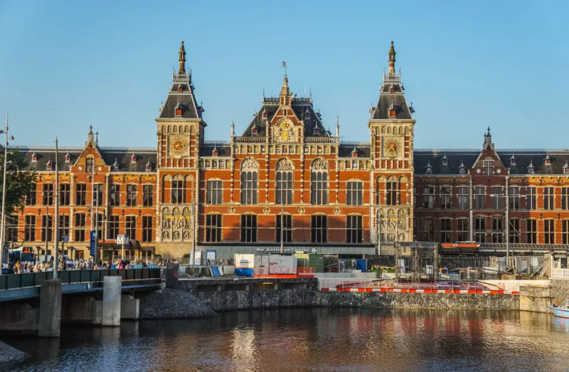 The iconic Amsterdam Central Station building captured in the warm glow of sunset with calm waters in the foreground – Who Should Actually Buy an Interrail or Eurail Pass?