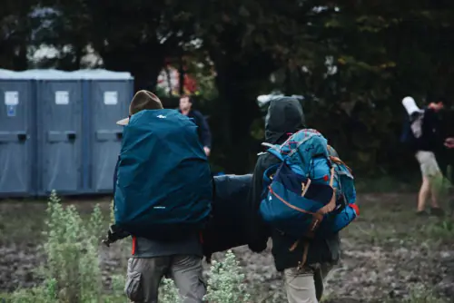 Two festival-goers carrying large backpacks past portable toilets at a muddy campsite – What Does Festival Travel Insurance Cover?